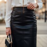 Žena v koženej sukni a bielej blúzke stojaca v mestskom prostredí za zlaté hodiny.